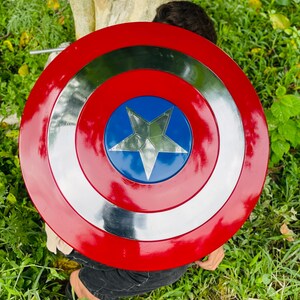 May include: A red, white, and blue shield with a silver star in the center. The shield has a circular design with concentric rings of red, silver, and blue. The shield is a prop or toy.