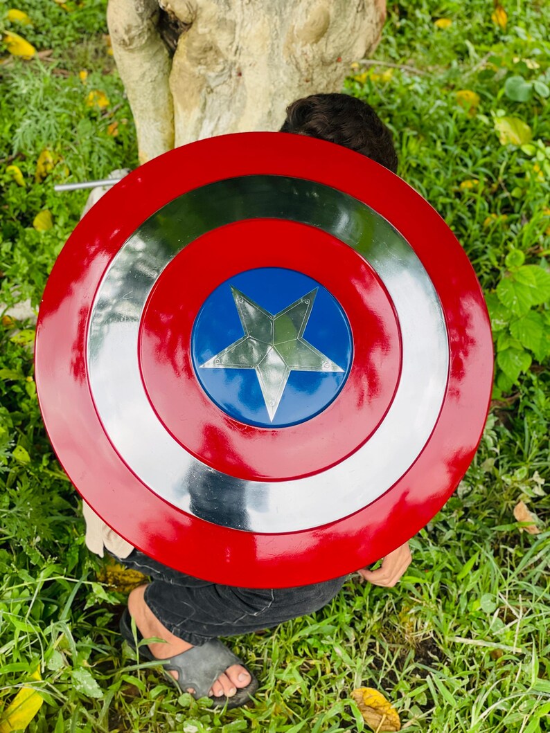 May include: A red, white, and blue shield with a silver star in the center. The shield has a circular design with concentric rings of red, silver, and blue. The shield is a prop or toy.