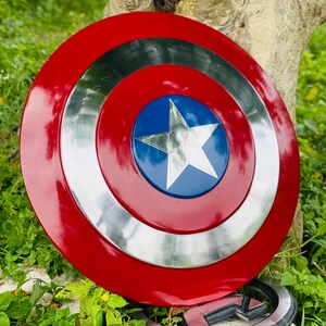 May include: A red, silver, and blue Captain America shield with a white star in the center. The shield is round with a glossy finish. A silver emblem is at the bottom of the image.