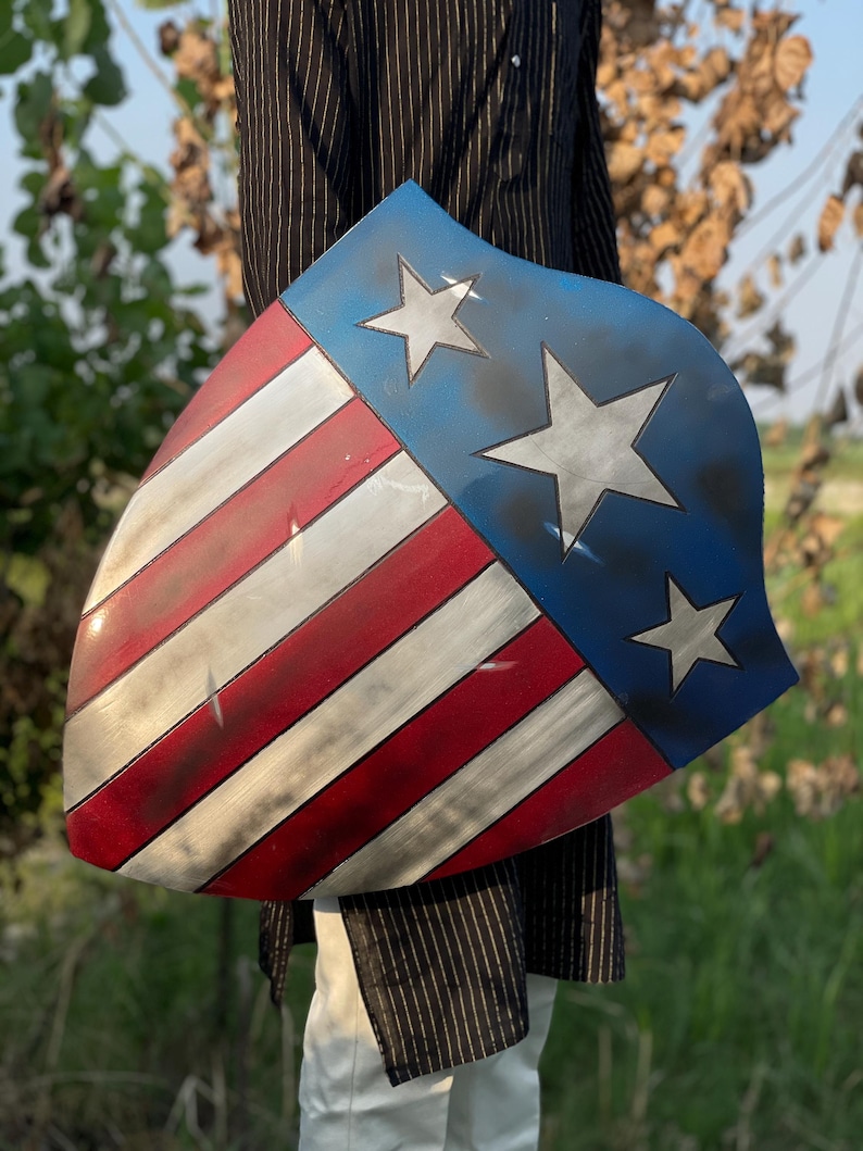Op de afbeelding: Een schild met een patriottisch ontwerp, met rode en witte strepen en een blauw gedeelte met drie witte sterren. Het schild heeft een verweerde, verouderde uitstraling, met zichtbare slijtage.
