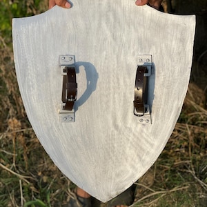 Op de afbeelding: Een zilver geschilderd houten schild met twee bruine leren riemen en metalen beugels aan de achterkant. Het schild heeft een gebogen bovenkant en een puntige onderkant. Het schild wordt door een persoon omhoog gehouden.