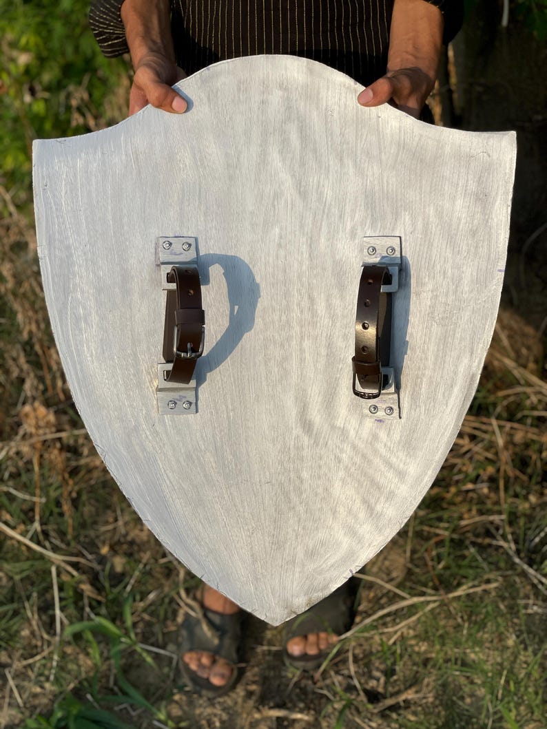 Op de afbeelding: Een zilver geschilderd houten schild met twee bruine leren riemen en metalen beugels aan de achterkant. Het schild heeft een gebogen bovenkant en een puntige onderkant. Het schild wordt door een persoon omhoog gehouden.
