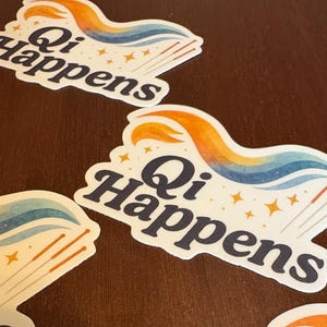 May include: White stickers with the text "Qi Happens" in a dark blue font. The stickers feature a colourful design with orange, blue, and yellow accents, along with gold stars and acupuncture needles. The stickers are on a brown surface.