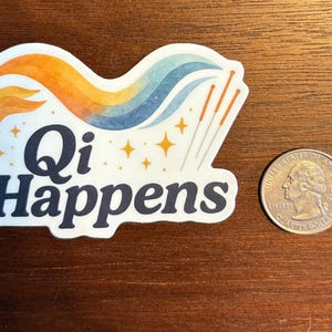 May include: A white sticker with the text "Qi Happens" in navy blue, featuring a colourful design of orange, yellow, and blue waves, with gold stars and acupuncture needles. A quarter coin is next to the sticker.
