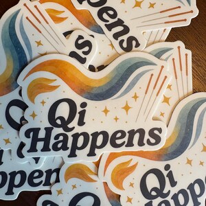 May include: A pile of white stickers with the text "Qi Happens" in black, stylish lettering. Each sticker has a colourful design with orange, blue, and yellow swirls, gold star accents, and acupuncture needles.