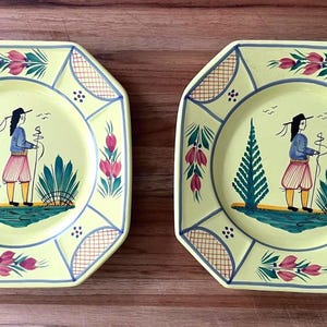 May include: Two octagonal decorative plates with a yellow background. Each plate features a central illustration of a figure in a blue jacket and pink shorts, holding a staff. The plates are adorned with floral and geometric patterns in blue, pink, and green.