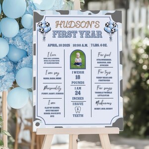 May include: A personalized sign for a baby's first year, featuring the text "HUDSON'S FIRST YEAR." The sign includes the baby's photo, weight (18 pounds), height (24 inches), and other milestones. The sign is decorated with blue and white accents.