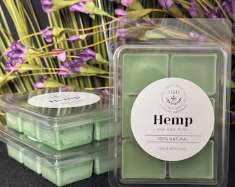May include: Green hemp soy wax melts in clear plastic packaging. The packaging includes a white label with the text "Hemp soy wax melt 100% natural made with love". The background features purple flowers.