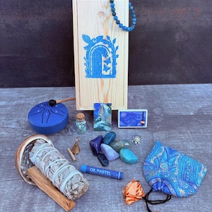 May include: A collection of spiritual items including a wooden box with a blue door design, a blue beaded bracelet, a blue tongue drum, crystals, sage, palo santo, and an oil pastel stick. The items are arranged on a wooden surface.