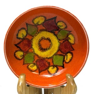 May include: Orange ceramic plate featuring a floral design in yellow, green, and red. The plate is displayed on a wooden stand. The plate's rim is a solid orange colour, and the floral design is in the centre.