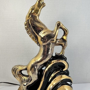 May include: A gold and black ceramic horse figurine in a rearing pose. The horse has a textured, metallic finish. The base is black with gold wave-like accents. A power cord extends from the base.