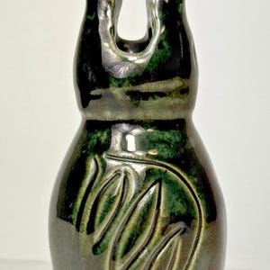 May include: A handcrafted ceramic vase with a glossy, dark green glaze. The vase has a unique, organic shape with a carved leaf design on the front. The top has a distinctive, open design.