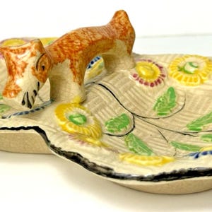 May include: A ceramic dish featuring a dog figurine. The beige dish has colourful floral patterns and a black rim. The dog is brown and white. The dish is likely for serving snacks or as a decorative item.