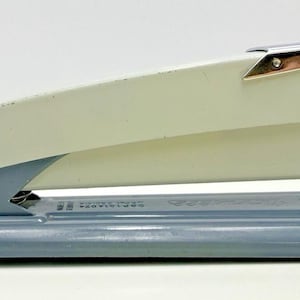 May include: A desktop stapler featuring a two-tone design with a cream-coloured top arm and a slate blue base. The stapler includes a metallic silver finish on the front tip and a silver-coloured metal plate at the front of the base for stapling.