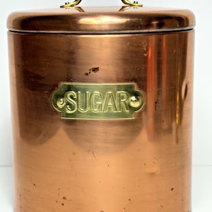 May include: A copper sugar canister with a brass handle and lid. The canister features a brass label reading "SUGAR". The copper has a reddish-brown hue, and the canister is cylindrical in shape.