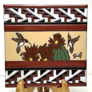 Peut inclure: Carreau de céramique carré avec un motif de colibri et de fleurs. La tuile présente un fond crème avec des fleurs marron, vertes et jaunes et deux colibris. La bordure présente un motif géométrique en blanc, noir et marron.