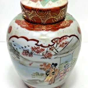 May include: A decorative ceramic jar with a lid. The jar features a detailed Japanese landscape scene with figures, trees, and a mountain. The lid and base have red and gold accents. The jar is approximately 13 cm tall.