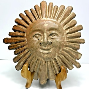 May include: A ceramic sun face sculpture with a smiling expression. The sun has a textured, tan surface with radiating rays. The sculpture is displayed on a wooden easel, showcasing its decorative nature.