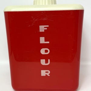 May include: A vintage red flour canister with a cream-coloured lid and a matching knob. The word "FLOUR" is printed vertically in white block letters on the front. The canister is rectangular with slightly tapered sides. Made for the kitchen.