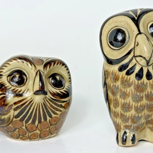 May include: Two ceramic owl ornaments. One is a rounded shape, the other is upright. Both have tan, brown, and black colouring. The owls have large, dark eyes and detailed feather patterns. The ornaments are decorative.