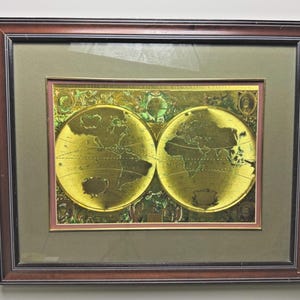 May include: Framed vintage map print showing a gold-toned world map with detailed continents and oceans. The map is set within a brown wooden frame, with a green mount and a pink inner border. The map is a historical cartographic representation.
