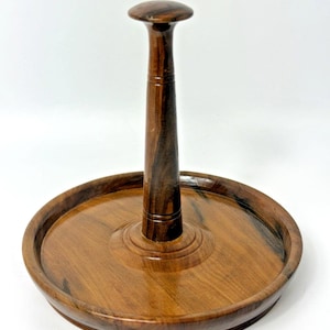May include: A handcrafted wooden cake stand with a dark brown finish. The stand features a round base with a raised edge and a central vertical support topped with a rounded handle. The wood grain is visible.