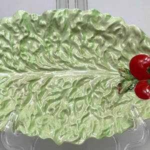 May include: A ceramic serving platter shaped like a lettuce leaf, with a light green colour and textured surface. Two red tomato decorations are on the right side of the platter. The platter is on a clear stand.