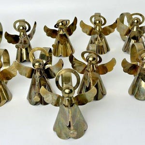 May include: A group of brass-coloured angel ornaments, each with wings and a halo. Some angels hold trumpets. The angels are displayed on a white surface, possibly for decoration or collection. The metal angels are likely vintage or antique.