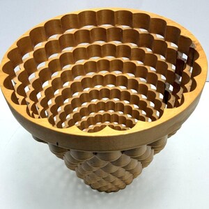 May include: A light brown wooden basket with a wide, circular opening and a tapered base. The structure is composed of interlocking, scalloped wooden rings that create a repeating, layered pattern throughout the entire body of the object.