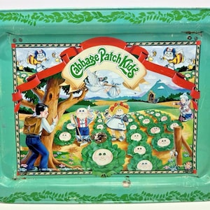 Peut inclure: Un plateau de service rectangulaire en métal avec une bordure vert clair ornée d'un motif de feuilles. Le centre présente une illustration Cabbage Patch Kids avec un jardin de choux, un arbre, une cigogne et des personnages, incluant le texte Cabbage Patch Kids.