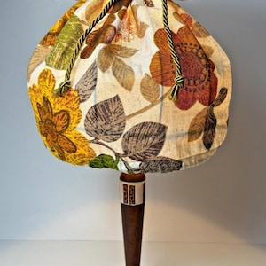Peut inclure: Lampe de table vintage avec un abat-jour à motifs floraux dans les tons de jaune, orange et marron. L'abat-jour est rassemblé en haut avec un cordon vert et or. La lampe a un pied en bois foncé et une base ronde en métal argenté.