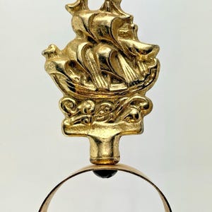 May include: A gold-coloured metal finial featuring a relief sculpture of a sailing ship with multiple masts and sails. The ship rests upon a decorative base with scrollwork details, and the entire piece is attached to a circular metal ring.