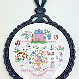 Peut inclure: Un dessous de plat rond en céramique avec un cadre en fonte noire et une poignée. Le centre blanc présente une illustration colorée d'une maison rose, d'un arbre, d'une vache, de moutons, de poules, d'un chat, d'un lapin, d'un oiseau et d'une personne dans une clôture rose.