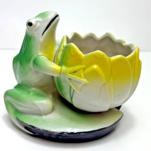 May include: A ceramic frog figurine in shades of green and white, holding a yellow and white flower-shaped container. The frog sits on a green lily pad base with a black rim. The container has a textured interior.