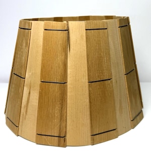 May include: A wooden lampshade with a trapezoidal shape. The shade is made from vertical wooden slats in varying shades of light brown and tan, joined by thin black lines. The design is simple and natural.