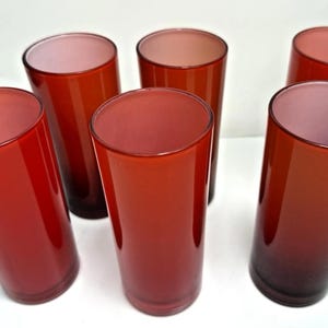 May include: Six tall, cylindrical drinking glasses in shades of red. The glasses have a gradient effect, transitioning from a lighter red at the top to a darker red at the base. The interior of each glass is a lighter pink.