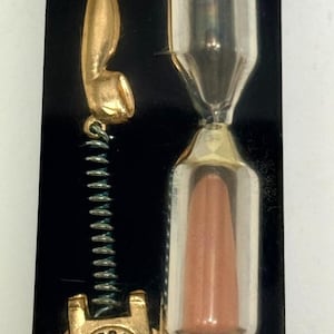 Peut inclure: Un objet rectangulaire noir présentant un téléphone à cadran doré avec un cordon en spirale et un sablier en verre transparent rempli de sable brun clair. Le combiné du téléphone est placé au-dessus de la base et le sablier est à côté.