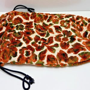 Peut inclure: Une pochette rectangulaire à cordon de serrage présentant un motif floral texturé dans des tons orange brûlé et vert olive sur un fond blanc cassé. Le sac est doté d'une doublure intérieure orange unie et d'une fermeture à cordon noir.