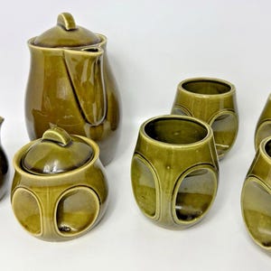 May include: An olive green ceramic tea set, comprising a teapot, creamer, lidded sugar bowl, and four distinctive cups with circular cutouts. The set's glossy finish and rounded forms evoke a retro style. The cups are approximately 8 cm in height.