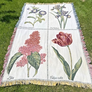 Peut inclure: Un plaid tissé présentant une grille de quatre illustrations botaniques. Les panneaux affichent un Convolvulus tricolor, un Iris xiphium, un lilas et une Tulipa culta avec leurs noms imprimés sous chaque fleur. Le plaid a un bord frangé.