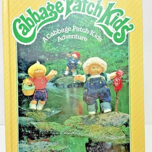 May include: A vintage "Cabbage Patch Kids" photo story book. The cover features the title in a green and white design, with illustrations of the dolls near a stream. The book has a yellow border with diagonal lines.