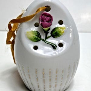 Peut inclure: Un ornement en céramique blanche en forme d'œuf avec une rose violette en relief et des feuilles vertes, de petits trous de ventilation circulaires et des lignes verticales dorées à la base, suspendu par un fin ruban doré.