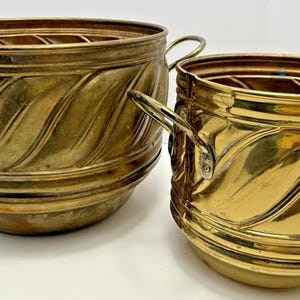 May include: Two round brass planters with a polished metallic finish and a diagonal ribbed texture. Each container features two curved side handles and a wide, open top. The brass displays natural variations in colour and tone throughout.