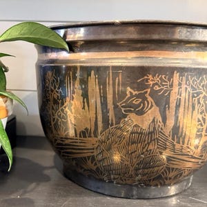 Peut inclure: Un cache-pot rond en métal avec une finition sombre et ternie, orné d'un motif gravé de tigre doré entouré d'arbres et de feuillages stylisés. Ce récipient en forme de bol présente un bord large et lisse ainsi qu'une base effilée.