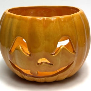 May include: A ceramic jack-o'-lantern with a glossy orange finish. The pumpkin has a carved face with triangular eyes, a nose, and a smiling mouth. The interior is hollow, and the pumpkin is designed to hold a candle.