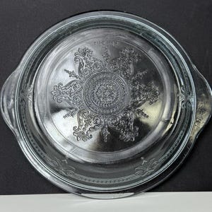 May include: A clear glass Fire King baking dish lid with a decorative floral pattern in the centre. The lid has two handles and the words "FIRE KING" are embossed on the rim. The dish is round and has a slightly raised edge.