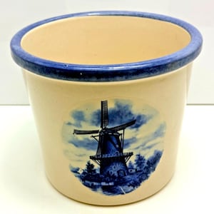 May include: A cream-coloured ceramic crock featuring a blue rim and a circular blue and white transfer print of a traditional windmill. The cylindrical vessel has a smooth, glazed finish and a wide opening, showcasing a classic design.