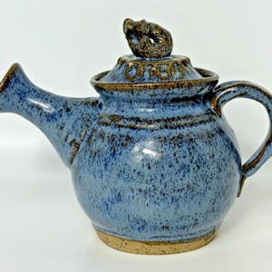 May include: A handmade ceramic teapot with a blue speckled glaze and a brown rim. The teapot has a curved spout, a rounded body, and a handle. The lid is topped with a decorative knob. The teapot sits on a tan base.