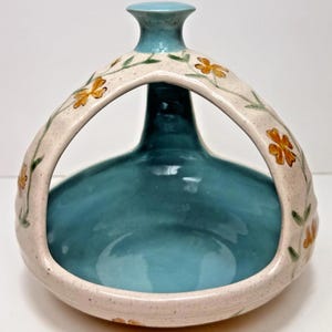 May include: A teal ceramic vase with a light blue neck and a cream-coloured handle. The handle is decorated with orange flower and green leaf patterns. The vase has a round base and a narrow opening.