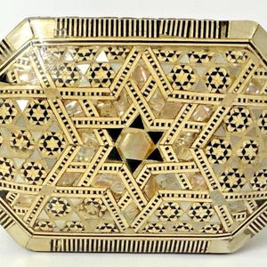 May include: An octagonal decorative box with an intricate inlaid design. The box features a central star pattern with black and white geometric shapes, surrounded by mother-of-pearl and ivory-coloured accents. The edges are lined with vertical black and white stripes.